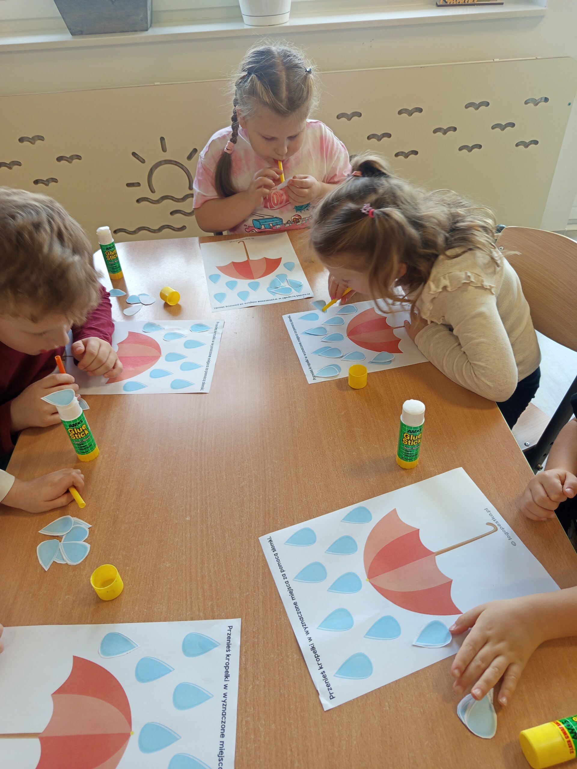 Dzieci przenoszą za pomocą słomki kropelki deszczu na parasol.